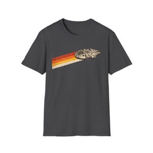 Puede incluir: Camiseta gris oscuro con un diseño retro de una nave espacial y una estela a rayas colorida. La nave espacial es beige y la estela es roja, naranja, amarilla y blanca. La camiseta es de cuello redondo.