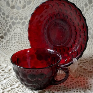 May include: A ruby red glass teacup and saucer set. The cup has a textured, bubble-like pattern, and the saucer has a scalloped edge. The set is displayed on a white lace tablecloth.
