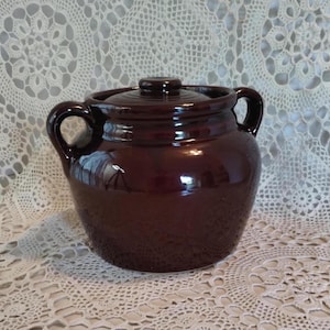May include: A dark brown ceramic bean pot with a glossy finish, featuring two side handles and a matching lid with a central knob. The pot has a rounded body and a wide opening, showcasing a smooth, reflective surface throughout the exterior.