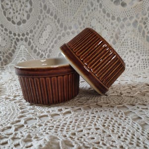May include: Two round ceramic ramekins with a glossy brown glaze and vertical ribbed detailing along the exterior sides. The interior of the ramekins is a smooth, light cream color, and the vessels feature a wide, open top design.