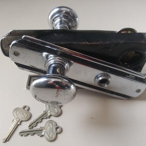 May include: A vintage chrome door handle with a black metal plate and three keys. The handle has a round knob and a lever.