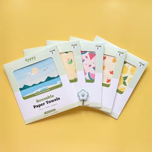 May include: A collection of reusable paper towels in various designs, including a landscape, watermelon, and lemons. The packaging features the word "Happy" and the text "Reusable Paper Towels". The towels are displayed on a yellow background.