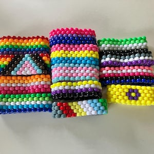 Könnte beinhalten: Eine Sammlung farbenfroher Perlenarmbänder in verschiedenen Pride-Flaggen-Designs. Die Armbänder zeigen Regenbogen-, Transgender-, Bisexualitäts- und Non-Binary-Flaggenmuster mit leuchtenden Farben wie Rot, Orange, Gelb, Grün, Blau und Lila.