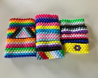 May include: A collection of colorful beaded bracelets in various pride flag designs. The bracelets feature a rainbow, transgender, bisexual, and non-binary flag patterns, with vibrant hues of red, orange, yellow, green, blue, and purple.