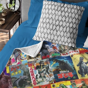 May include: A soft blanket with a collage of vintage Godzilla film posters in various colours. The blanket is on a bed with blue sheets and a patterned pillow. The blanket's design includes Japanese text.