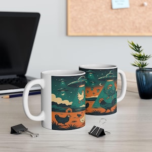 May include: Two white ceramic mugs with a whimsical design featuring chickens, UFOs, and a farm scene. The illustration includes a night sky, green spotlights, and an orange field. The mugs are on a light-colored wooden surface.
