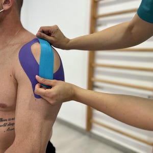 Könnte beinhalten: Schulter einer Person mit violettem und blauem Kinesiologie-Tape. Das Tape wird von einer anderen Person angebracht. Der Hintergrund umfasst eine Holzwand und eine weiße Wand.