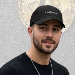 Puede incluir: Gorra de béisbol negra con el texto "I told you so" en blanco. La gorra la lleva una persona. Un gráfico de Bitcoin está en el fondo.