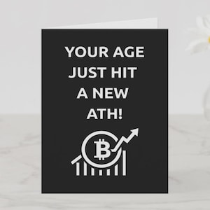 May include: A black greeting card with white text that reads "YOUR AGE JUST HIT A NEW ATH!". The card features a Bitcoin symbol with an upward trending arrow. A vase with a daisy is in the background.