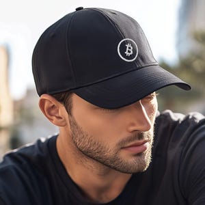 May include: A black baseball cap featuring a white embroidered Bitcoin symbol inside a circular outline on the front panel. The cap has a structured crown, a curved brim, and a solid black fabric construction with a button on the top center.