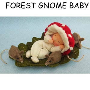 Peut inclure: Un bébé gnome en feutre portant un chapeau de champignon rouge et blanc, dormant sur une feuille de feutre verte avec trois souris en feutre brun.