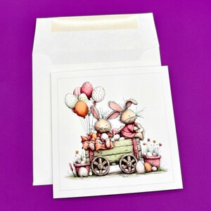 May include: Easter greeting card featuring two cartoon bunnies riding in a wooden cart filled with Easter eggs. The bunnies are holding balloons. The card is inside a white envelope, set against a purple background.