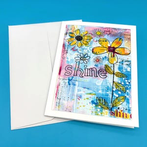 Op de afbeelding: Een wenskaart met een bloemenontwerp en het woord "shine". De kaart heeft gele en witte bloemen met groene bladeren tegen een blauwe en roze achtergrond. Een witte envelop is inbegrepen.