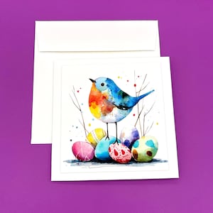 Pode incluir: Cartão de felicitações em aquarela com um pássaro colorido empoleirado em cima de um monte de ovos de Páscoa decorados. O pássaro tem plumagem azul, laranja e amarela. O cartão é branco com um envelope a condizer, sobre um fundo roxo.