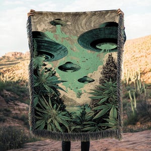 Peut inclure: Une couverture tissée avec une illustration rétro d'OVNIs survolant un paysage avec des plants de cannabis. La couverture a une bordure à franges et est présentée sur un fond désertique. La palette de couleurs comprend des nuances de vert, de bleu et de marron.