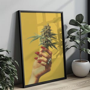 May include: A framed print featuring a hand with red nails holding a cannabis bud against a yellow background. The print is in a black frame and is leaning against a wall. A plant in a white pot is in the background.
