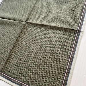 May include: A green and white patterned scarf with a chevron design. The scarf has a border with blue and pink stripes. The brand name "YVES SAINT LAURENT" is printed in white at the bottom right corner.