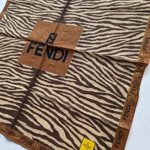 May include: A brown and tan zebra-striped scarf featuring the Fendi logo in the centre and along the border. The scarf is folded, revealing the brand name and logo in a square patch. The scarf has a tag with the Fendi logo.