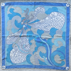 May include: A square silk scarf featuring a blue and grey graphic illustration of a figure with large, intricate dragonfly-like wings. The design includes swirling cloud patterns and the text Maersk Line printed in the corners of the border.