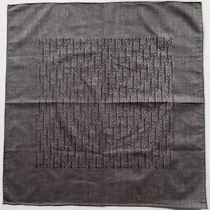 May include: A gray square scarf with a repeating pattern of the letters "D" and "i" in a darker gray shade. The scarf has a textured appearance and a subtle sheen.