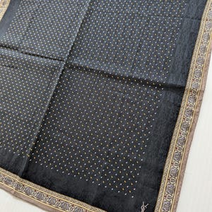 May include: Black silk scarf featuring a pattern of small white and gold dots. The scarf has a decorative border with a brown, gold, and red design. A silver logo is in the lower right corner.