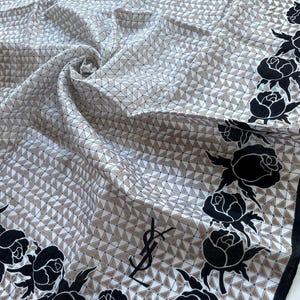 May include: A silk scarf with a geometric pattern of triangles in white and beige. Black rose illustrations border the edges. The scarf features the Yves Saint Laurent logo in black.
