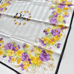 May include: A white silk scarf with a floral design featuring yellow and purple flowers. The scarf has a striped background, a black border, and the Christian Dior logo. The flowers are arranged in a decorative pattern.