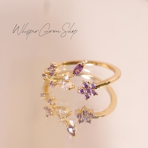 May include: A gold-colored ring with a floral design featuring purple and clear gemstones. The ring has a delicate, open design with a flower and leaf motif. The text "Whisper Grove Shop" is written in cursive.