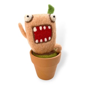 May include: A felt plant with a brown body, a red mouth, and white teeth. The plant has a green leaf on top and is in a brown pot.