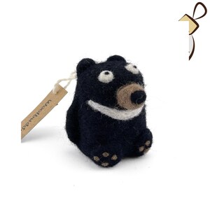 Puede incluir: Un oso de fieltro negro con una sonrisa blanca y una nariz de fieltro marr&oacute;n. El oso tiene ojos grandes y blancos y est&aacute; sentado sobre una superficie blanca. El oso tiene una etiqueta marr&oacute;n que dice "Woolbuddy".