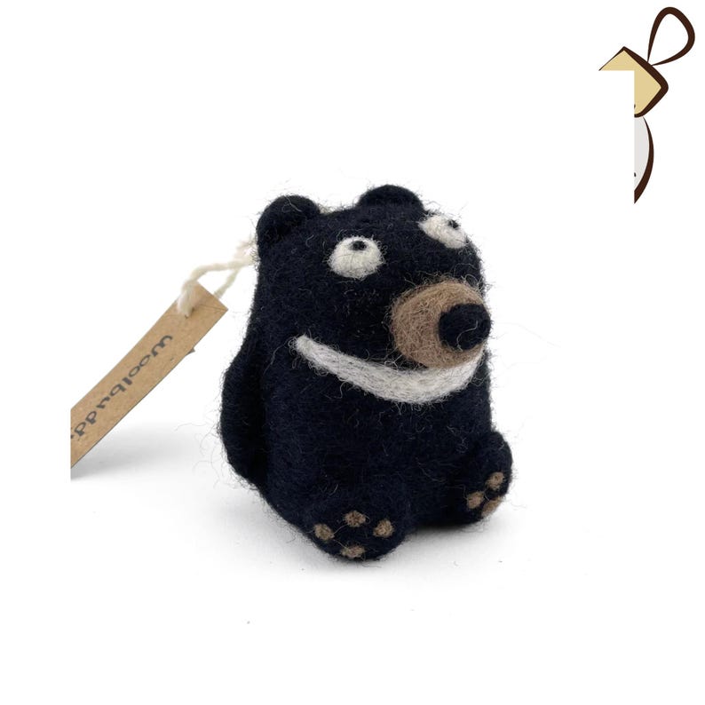 Puede incluir: Un oso de fieltro negro con una sonrisa blanca y una nariz de fieltro marr&oacute;n. El oso tiene ojos grandes y blancos y est&aacute; sentado sobre una superficie blanca. El oso tiene una etiqueta marr&oacute;n que dice "Woolbuddy".
