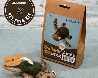 May include: A brown cardboard box with a cut-out handle contains a needle felting kit to make a sea turtle. The box has the text "Woolbuddy" and "Sea Turtle Kit Needle Felting" on it. A finished felt sea turtle is shown in front of the box.