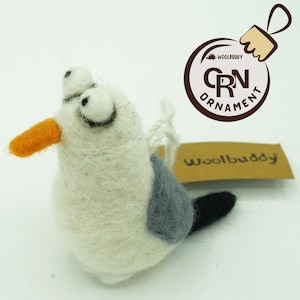 May include: A handmade felt seagull ornament with an orange beak, gray wing patch, and black tail. The seagull has large, expressive eyes and is attached to a brown tag that reads "woolbuddy". A brown circular ornament with the text "CRN ORNAMENT" is also visible.