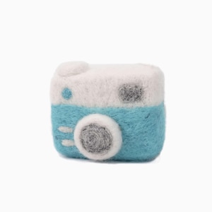 May include: A handmade felt camera in a square shape. The top is white, the bottom is turquoise, and the lens is gray. A small blue dot and a gray square are on the top. The camera has a round lens and two white lines on the front.