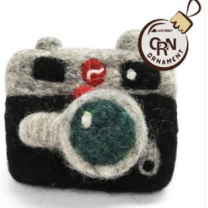 May include: A handmade felt camera ornament in gray, black, red, and green. The ornament has a lens, buttons, and the text "CRN Ornament" on a tag. The ornament is designed to hang on a Christmas tree.