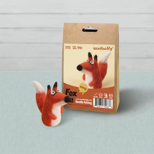 May include: A Woolbuddy needle felting craft kit in a brown paper bag with a fox illustration and the text Fox Kit, featuring a finished orange and white felted fox figurine with black ears and nose, designed for ages 14 and up.
