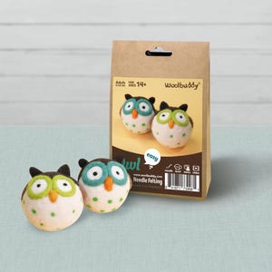 May include: A Woolbuddy needle felting craft kit in a brown paper bag featuring two finished felted owl figures. The owls are white with green polka dots, dark brown ear tufts, and bright green eye rings with orange beaks. The packaging includes text.