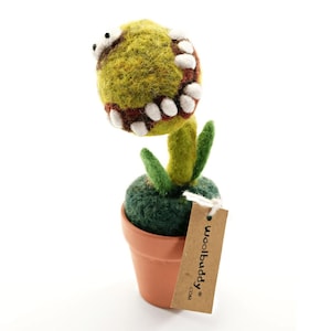May include: A handmade felt plant sculpture in a terracotta pot. The plant has a large, round, green head with white teeth and brown accents. The stem and leaves are green. A brown tag with the text "woolbuddy" hangs from the pot.