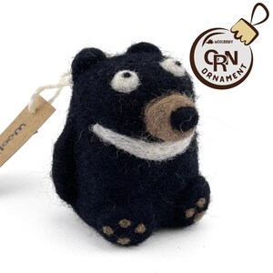 Puede incluir: Un adorno de oso de fieltro negro con una sonrisa blanca, nariz marr&oacute;n y ojos blancos. El adorno tiene una etiqueta de fieltro marr&oacute;n que dice "Wool" y una etiqueta de fieltro marr&oacute;n que dice "Woolbuddy Ornament CRN".