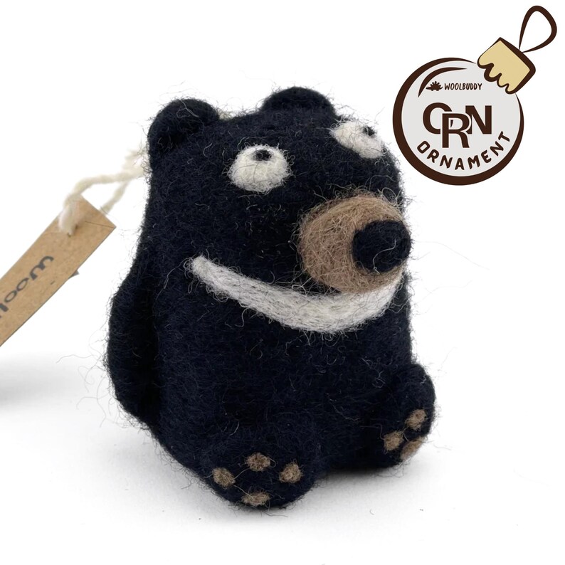 Puede incluir: Un adorno de oso de fieltro negro con una sonrisa blanca, nariz marr&oacute;n y ojos blancos. El adorno tiene una etiqueta de fieltro marr&oacute;n que dice "Wool" y una etiqueta de fieltro marr&oacute;n que dice "Woolbuddy Ornament CRN".
