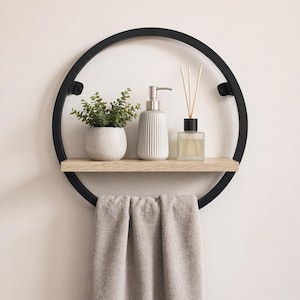 May include: A circular black metal wall shelf with a light wood shelf and a grey towel hanging below. The shelf holds a small potted plant, a white ribbed soap dispenser, and a glass diffuser with reeds.