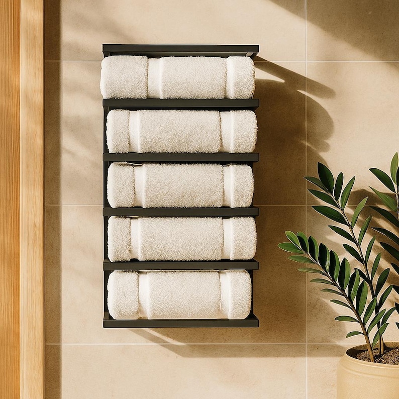 May include: A black wall-mounted towel rack with five shelves, each holding a rolled white towel. The towels are neatly arranged, creating a clean and organized look. The rack is mounted on a light-colored tiled wall, with a plant in a pot in the corner.