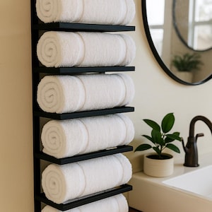 May include: A black metal towel rack with six shelves, each holding a rolled white towel. The rack is mounted on a light-colored wall, near a bathroom sink and mirror. The towels appear soft and fluffy.