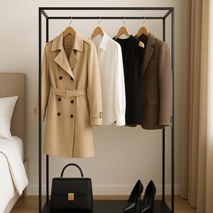 Op de afbeelding: Een zwart metalen kledingrek toont een beige trenchcoat, een wit overhemd, een zwarte top en een bruine blazer. Een zwarte handtas en zwarte pumps staan op de onderste plank. Een selectie van kleding.