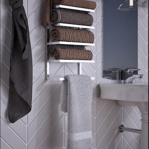May include: A modern bathroom scene featuring a chrome towel rack with rolled brown towels and a hanging gray towel. A dark gray towel hangs on the left. The background includes a mirror, sink, and gray tile.