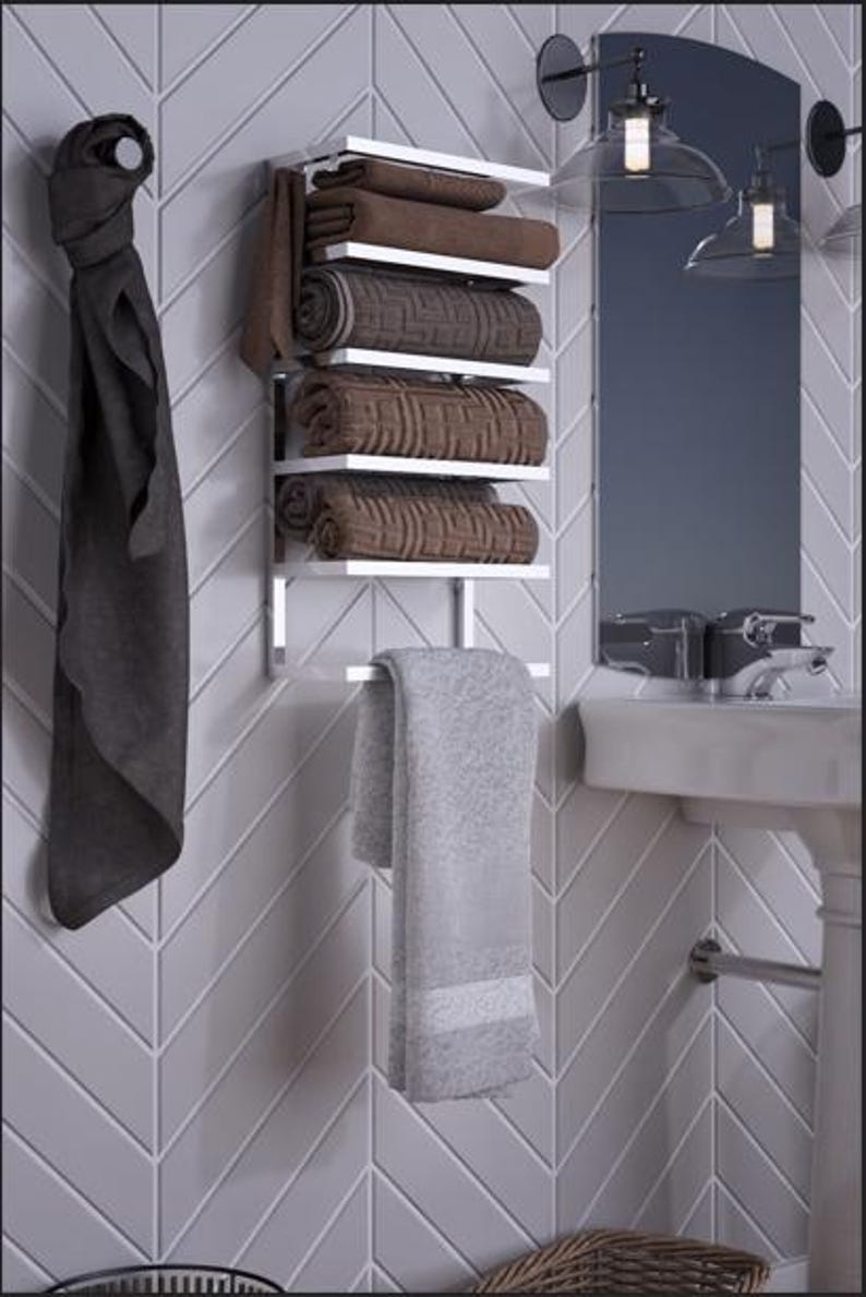 May include: A modern bathroom scene featuring a chrome towel rack with rolled brown towels and a hanging gray towel. A dark gray towel hangs on the left. The background includes a mirror, sink, and gray tile.