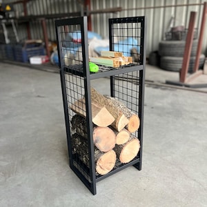 May include: A black metal log store with a grid design. The rack is filled with cut logs and has a top shelf with small wooden pieces and a green object. The rack is in an industrial setting.