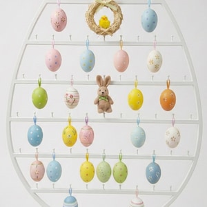 May include: White metal Easter egg display with hanging pastel eggs, a small bunny, and a wreath with a yellow chick. A cluster of eggs and daffodils are at the base.