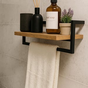 Peut inclure: Étagère de salle de bain moderne avec cadre en métal noir et étagère en bois. Elle contient une bouteille de savon pour les mains marron, une petite plante en pot, un diffuseur noir et une tasse noire. Une serviette blanche est suspendue en dessous.