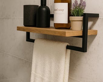 May include: A modern bathroom shelf with a black metal frame and a wooden shelf. It holds a brown hand soap bottle, a small potted plant, a black diffuser, and a black cup. A white towel hangs below.